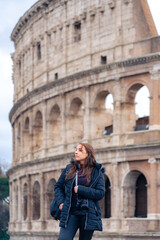 Fototapeta na wymiar Pensive young Latin woman tourist in warm clothes and professional camera standing against Colosseum and looking away while admiring historic architecture during trip through Rome, Italy