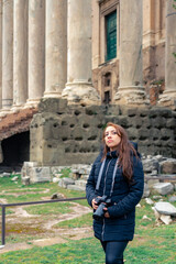 Fototapeta na wymiar Pensive young Latin woman tourist in warm clothes and professional camera standing in Roman Forum next to Colosseum and looking away while admiring historic architecture during trip in Rome, Italy