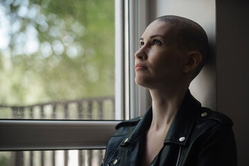 Beautiful young woman with a shaved head without hair sits and looks out the window