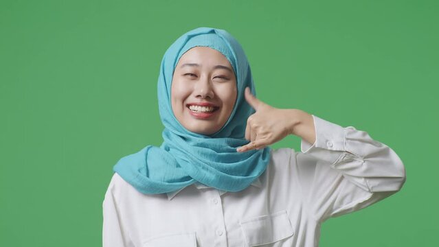 Close Up Of Asian Muslim Woman Smiling And Making Call Me Gesture While Standing In The Green Screen Background Studio
