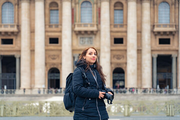 Fototapeta premium Full body of young Latin woman tourist in trendy clothes with professional photo camera standing in front of historic church Saint Peter Basilica in Vatican city, Rome, Itay