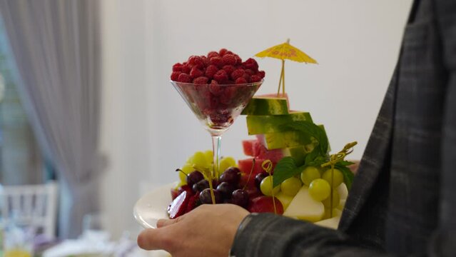 Man in business suit holds tray of sliced and beautifully arranged fruits and raspberries in martini glass. In banquet hall of restaurant, head brought compliment as an apology to guest.