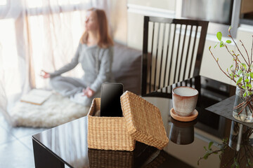 Smartphone is in separate box on table. Woman meditating at window in background. Stop using digital gadgets at home in favor of reading books and meditation. Mental health and digital detox concept