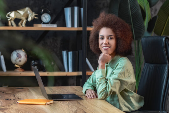 Portrait of happy confident African American businesswoman. Successful female entrepreneur sitting at the workplace at the table with a laptop in office looking confident at the camera and smiling