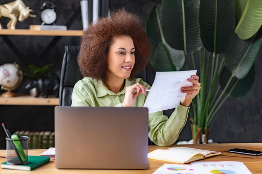 Smiling business woman, company employee sitting at the workplace in the office analyzes documents, focused on looking through report, sales schedule