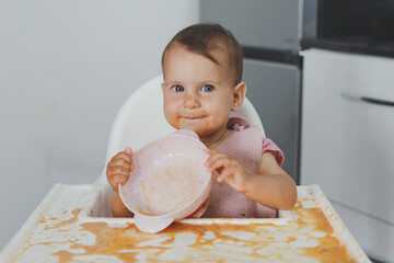 funny cheerful dirty baby sits in a highchair in the kitchen and eats complementary foods, holds an...