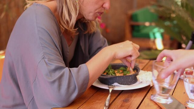 Stylish woman having breakfast in summer cafe terrace, side view of a hungry woman dipping bread into pan with delicious Georgian shakshuka omlette