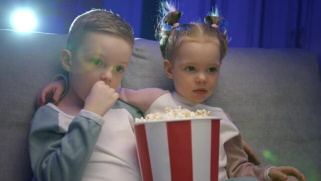 Brother and sister eat popcorn from bucket staring at image from movie projector