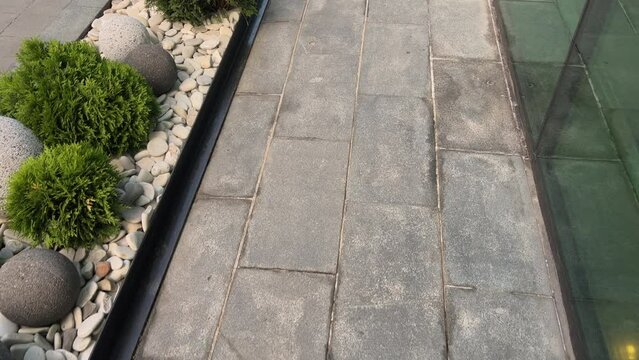 POV of a person walking on tiles past a glass facade and flowerbed decorated with luminaires, rocks and bushes