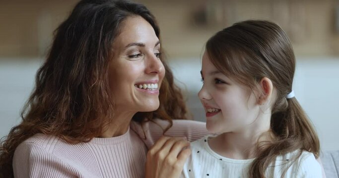 Little daughter whispers secrets on mothers ear, telling confidential information, gossiping together sit on sofa at home. Trust and understanding, trustworthy relationships between parent and child
