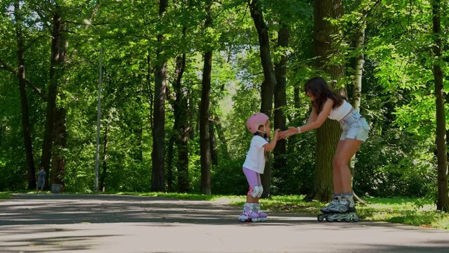 Mom teach little daughter girl inline skating with roller in public park in summer sunny day. Happy family time together, Leisure sport activity.