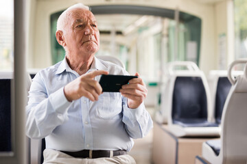 Senior European man sitting on seat inside tram and using his smartphone.