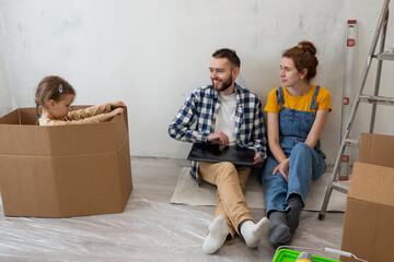 Fototapeta na wymiar Family sit on floor in new apartment during renovation process with laptop. Planning for furniture in new home. Online shopping or house decor together.