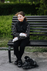 Fototapeta premium A girl in sadness, a child athlete with a cast, a bandage on her hands after a bone fracture, sits on a bench in the park with roller skates, missing riding alone. Photography, portrait, sports.