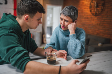 Fototapeta na wymiar teenage boy and mature caucasian woman sit together at the kitchen at home talk mother and son or relatives support solving problem share experience and opinion boy hold mobile phone smartphone