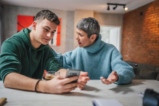 teenage boy and mature caucasian woman sit together at the kitchen at home talk mother and son or relatives support solving problem share experience and opinion boy hold mobile phone smartphone