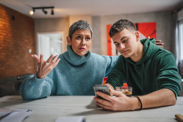 teenage boy and mature caucasian woman sit together at the kitchen at home talk mother and son or relatives support solving problem share experience and opinion boy hold mobile phone smartphone