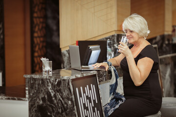 Attractive adult woman sitting at bar counter with laptop and drinking water