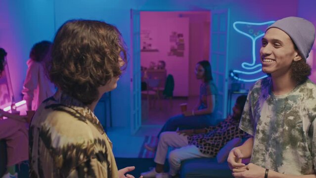 Medium shot of young Arab man in beanie and tiedye tshirt chatting to male friend with curly hair at house party with blue and pink neon lighting, and other guests talking in background
