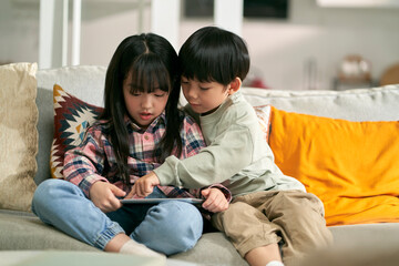two cute little asian kids brother and sister sitting on family couch at home playing game using...