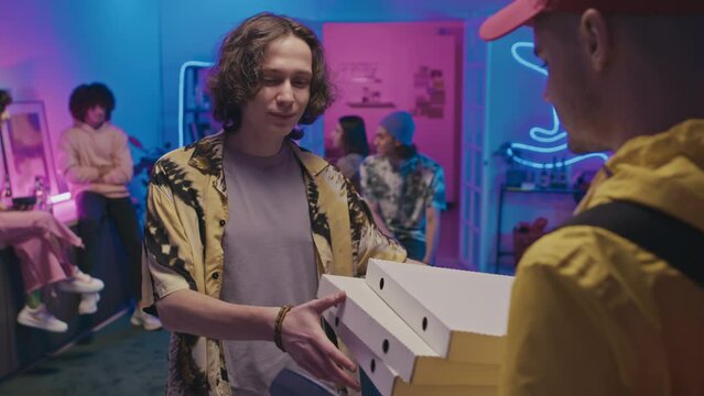 Medium shot of young Caucasian guy talking to delivery courier at house party, paying by scanning smart watch on wireless terminal, then carrying pizza over to friends, and everybody cheering