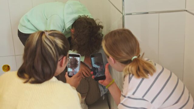 Teenage girls recording video on their smartphones of their classmate being bullied in school corridor by another boy teasing him