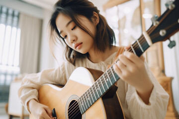 Fototapeta premium Beautiful, charming Japanese girl playing, practicing acoustic guitar in her cozy bedroom with concentration in natural light from window