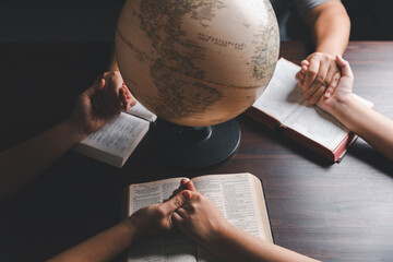 Christian group praying for globe and people around the world on wooden table with bible. Christian small group holding hands and praying together around a wooden table with bible page in homeroom. - obrazy, fototapety, plakaty
