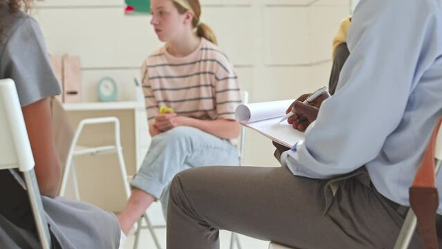 Cropped shot of unrecognizable Black male school psychologist taking notes on clipboard while listening to multiethnic kids sharing their mental problems during group therapy session