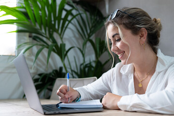 Millennial smiling caucasian girl sit at desk in living room study on laptop making notes, concentrated young woman work on computer write in notebook, take online course or training at home education