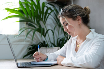 Millennial caucasian girl sit at desk in living room study on laptop making notes, concentrated young woman work on computer write in notebook, take online course or training at home, education 