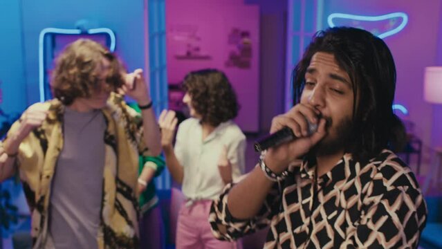 Medium shot of young Middle Eastern guy with beard and long hair singing into microphone at house party, in pink and blue neon light, and male and female friends dancing energetically to rhythm