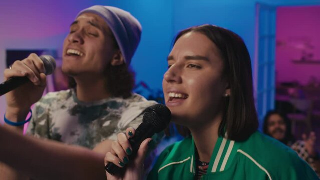 Close-up shot of young artistic multiracial couple singing karaoke song into microphone together at student party, in room with neon lighting, and friends dancing and clapping in background