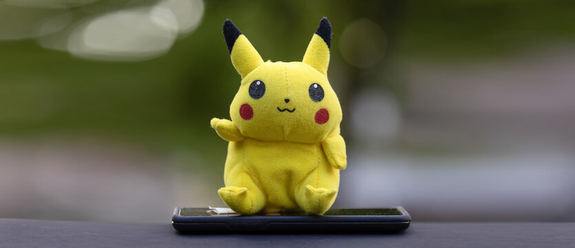 Editorial Content, Plush Pikachu.  Image of a stuffed, plush yellow Pikachu from Pokemon  sitting on top of a smart phone, outside.  Editorial.