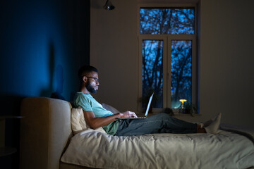Focused African American programmer man in dark room lying on bed work on laptop. Workaholic freelancer black guy sitting comfortably with computer on laps working on graphic design for app at night