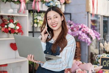 One young Asian female florist owner, working with laptop, selling floral arrange, talking on mobile phone in colorful flower shop store with a beautiful bunch of blossoms, and e-commerce business. - obrazy, fototapety, plakaty