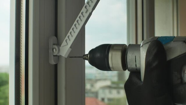 Male uses electric drill to drill holes in plastic window frame to install window stopper. Drilling hole for fasteners to install window limiter, close-up. Drilling with electric drill. Home repairs