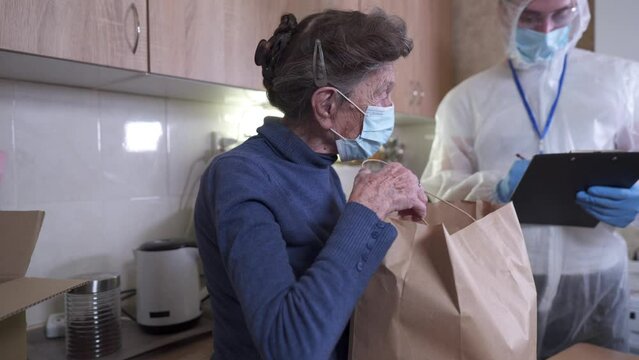 Male social worker of organization helping lonely old people places food donations on table at senior woman house. Volunteer in protective suit from food bank. Happy elderly female food gets endowment