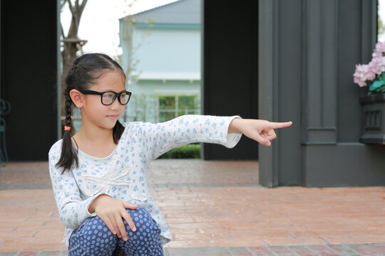Asian young girl kid in glasses pointing index finger to beside while sitting in the pavilion at the garden.