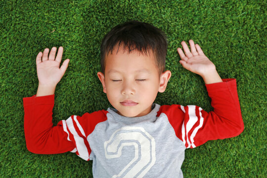 Peaceful Asian little boy close eyes and lying down on green lawn. Kid lies on grass. Top View.