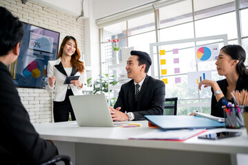 A cheerful and confident Asian businesswoman stands, present bar charts data from a whiteboard to her office colleagues. Asian business women leader role at the meeting.