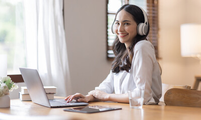 Young adult happy smiling Hispanic Asian student wearing headphones talking on online chat meeting using laptop in home office. College female student learning business education remotely.