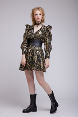 High fashion photo of beautiful elegant young woman in a pretty military camouflage dress with sleeves, boots, wide belt posing over white, soft gray background. Studio Shot, portrait. Blonde