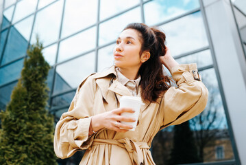 Stylish mixed race woman in raincoat holding disposable cup of coffee fixing hair while standing against modern glass building background, urban lifestyle