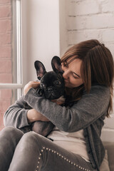 A young woman is embracing a puppy, a black French Bulldog, sitting on the windowsill at home.The concept of care, training,raising of animals.