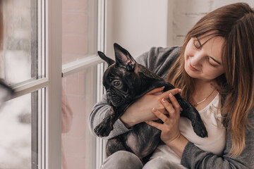 A young woman is embracing a puppy, a black French Bulldog, sitting on the windowsill at home.The concept of care, training,raising of animals.