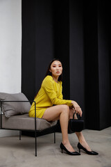 High fashion photo of beautiful elegant young asian woman in pretty yellow short dress, handbag, clutch posing on textured black and white wall. Slim figure. Model sits on chair