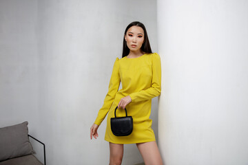High fashion photo of a beautiful elegant young asian woman in pretty short yellow dress, handbag, clutch. White textured rounded wall