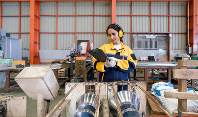 Young female labor dressed in polyester jacket uniform and ear muff reading document on clipboard, stand in front of steel and metal processing machinery. Atmosphere in metal sheet factory.