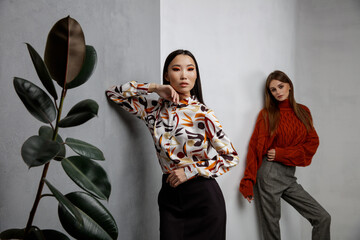 Two fashion models in orange beige looks, outfits. Sweater, shirt, long black skirt, gray pants, trousers. Beautiful young women.  Asian and european girls posing on textured wall, tall flower